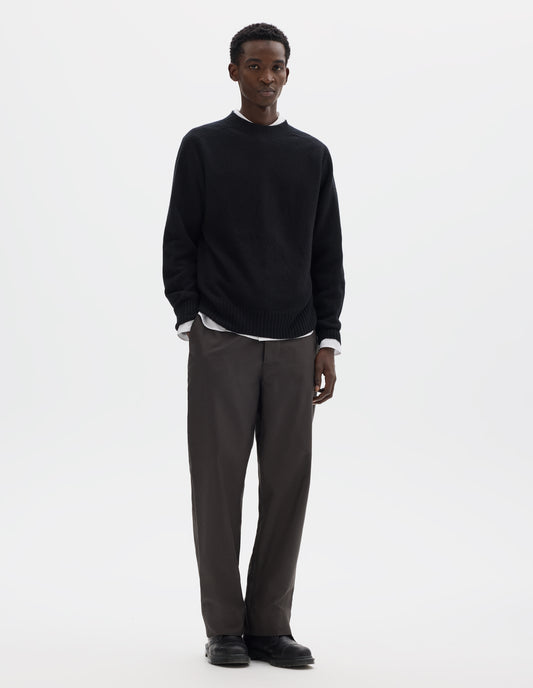 Full body image, front view of model wearing black merino cashmere seamless jumper with ribbed crew neck, cuffs and hem. Styled over white shirt, and with grey trousers and black leather shoes.