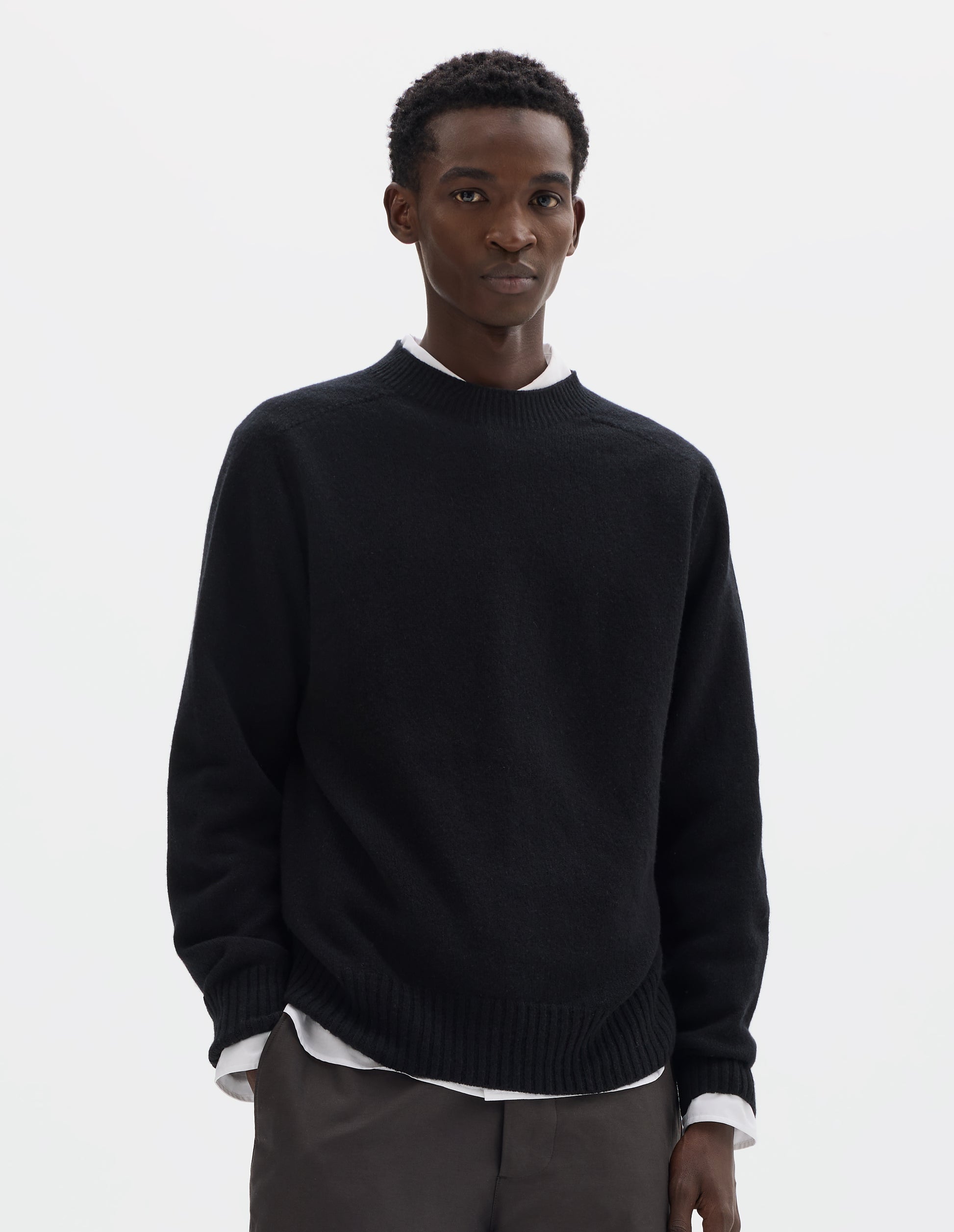Cropped body image, front view of model wearing black merino cashmere seamless jumper with ribbed crew neck, cuffs and hem. Styled over white shirt and grey trousers.