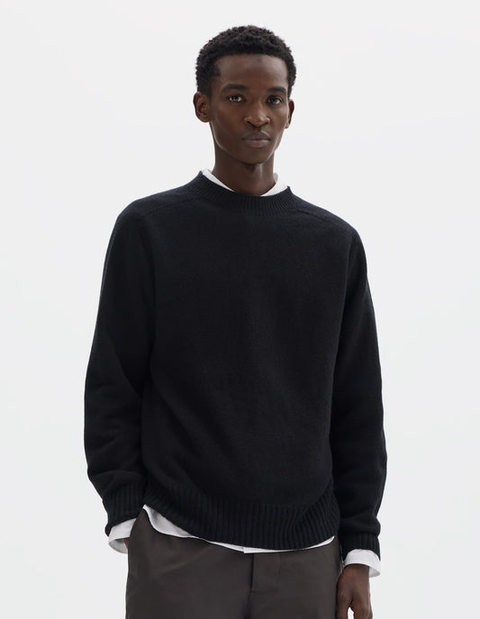 Cropped body image, front view of model wearing black merino cashmere seamless jumper with ribbed crew neck, cuffs and hem. Styled over white shirt and grey trousers.