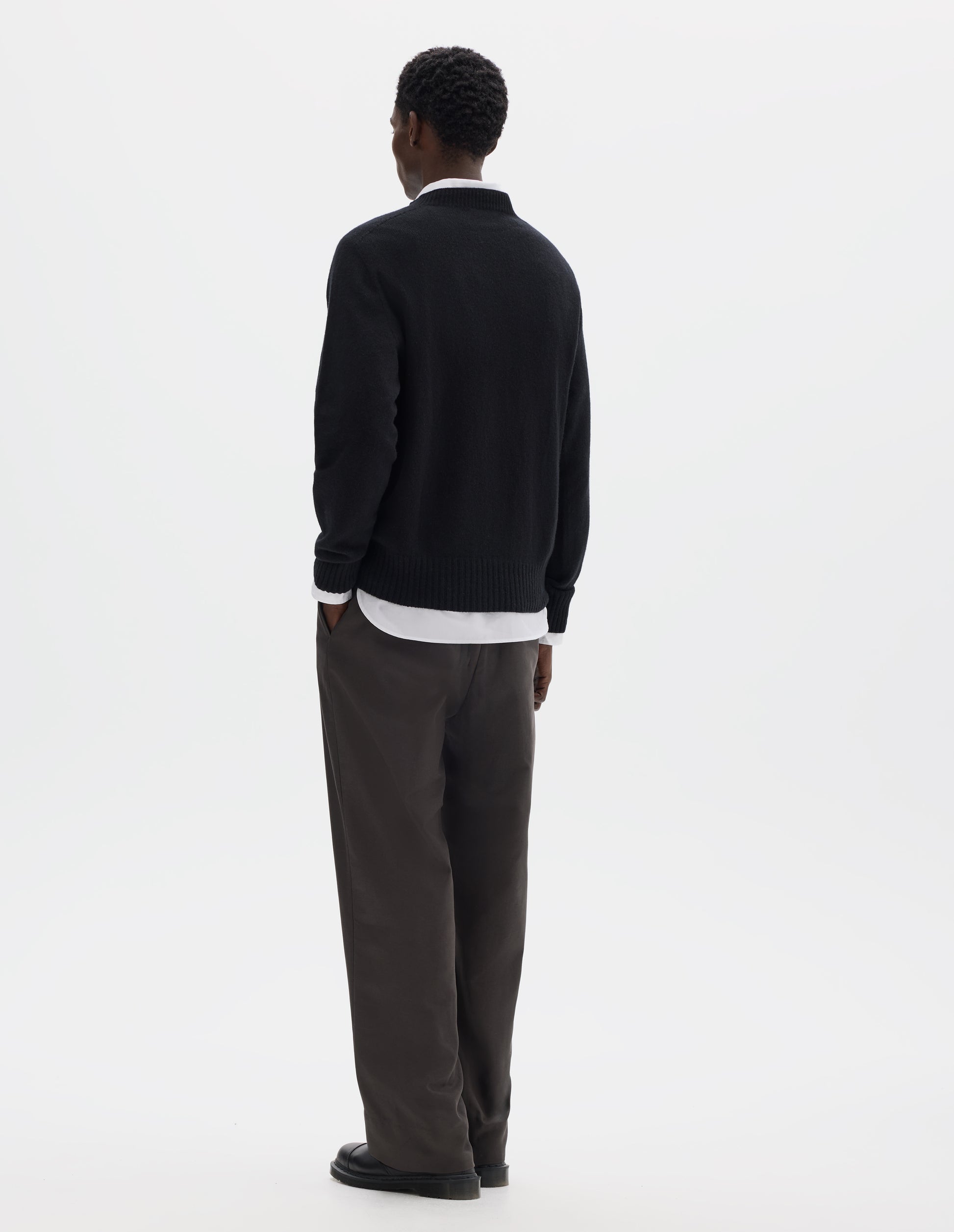Full body image, back view of model wearing black merino cashmere seamless jumper with ribbed crew neck, cuffs and hem. Styled over white shirt, and with grey trousers and black leather shoes.