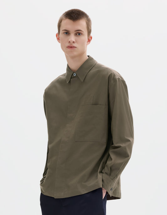 Cropped body image, front view of model wearing green relaxed cotton shirt with concealed placket and mother of pearl button on collar, large patch chest pocket with internal pen pocket and vent split hem. Styled with dark blue trousers.
