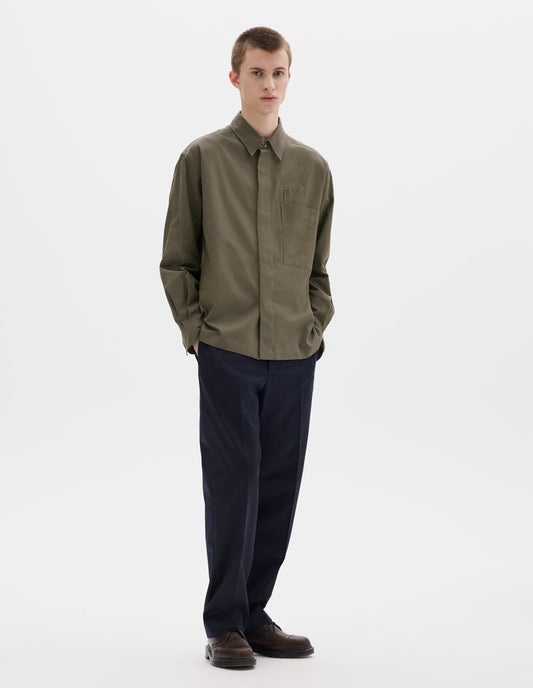 Full body image, front view of model wearing green relaxed cotton shirt with concealed placket and mother of pearl button on collar, large patch chest pocket with internal pen pocket and vent split hem. Styled with dark blue trousers and brown leather shoes.