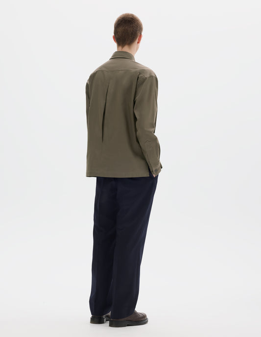 Full body image, back view of model wearing green relaxed cotton shirt with mother of pearl buttons on cuffs, deep inverted pleat at back yoke and vent split hem. Styled with dark blue trousers and brown leather shoes.