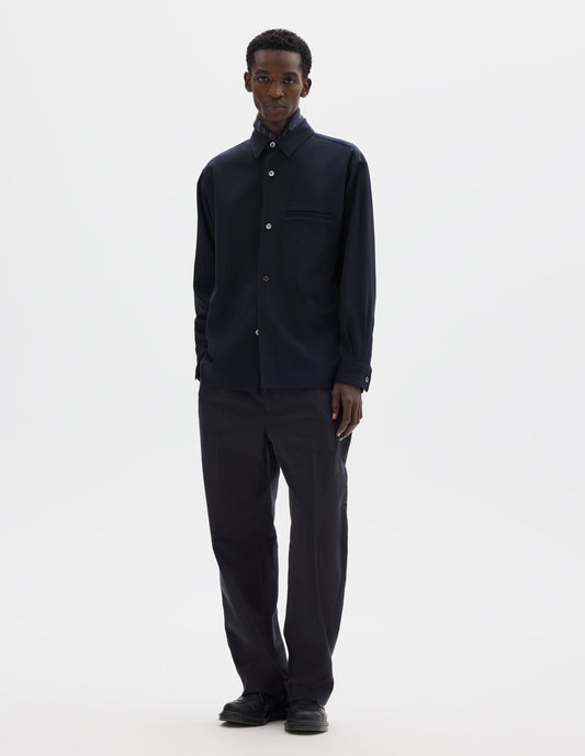 Full body image, front view of model wearing relaxed dark blue wool shirt with mother of pearl buttons and welt chest pocket. Styled with dark blue silk scarf, black trousers and black leather shoes.