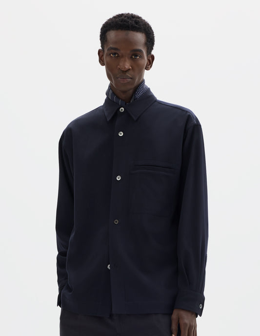 Cropped image, front view of model wearing relaxed dark blue wool shirt with mother of pearl buttons and welt chest pocket. Styled with dark blue silk scarf and black trousers.