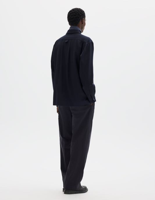 Full body image, back view of model wearing relaxed dark blue wool shirt with mother of pearl buttons on cuffs, wide box pleat and hanger loop detail on back yoke. Styled with dark blue silk scarf, black trousers and black leather shoes.
