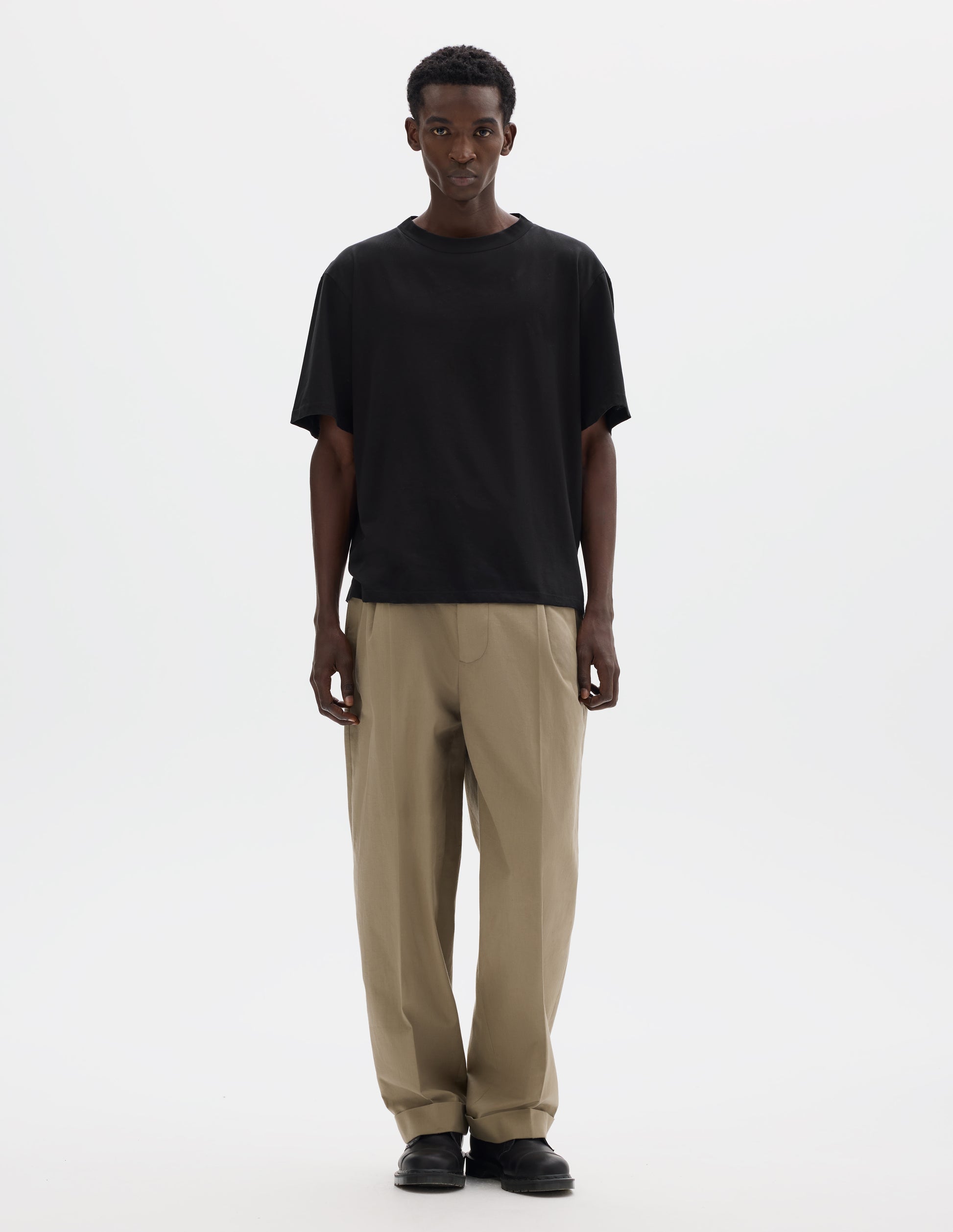Full body image, front view of model wearing black relaxed cotton t-shirt with crew neck and boxy cut. Styled with beige trousers and black leather shoes.
