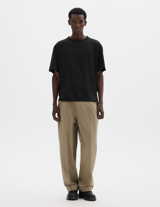 Full body image, front view of model wearing black relaxed cotton t-shirt with crew neck and boxy cut. Styled with beige trousers and black leather shoes.