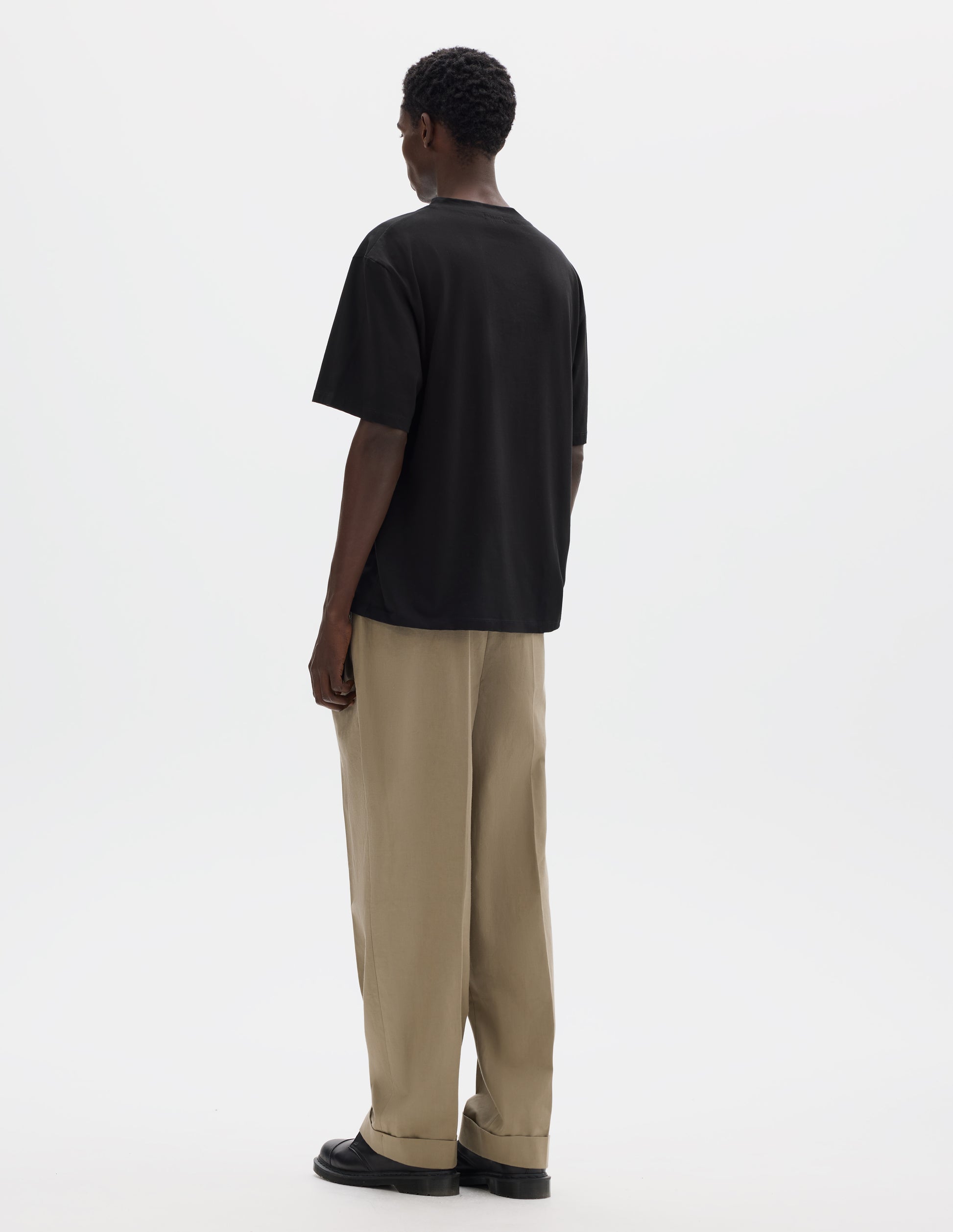 Full body image, back view of model wearing black relaxed cotton t-shirt with crew neck and boxy cut. Styled with beige trousers and black leather shoes.