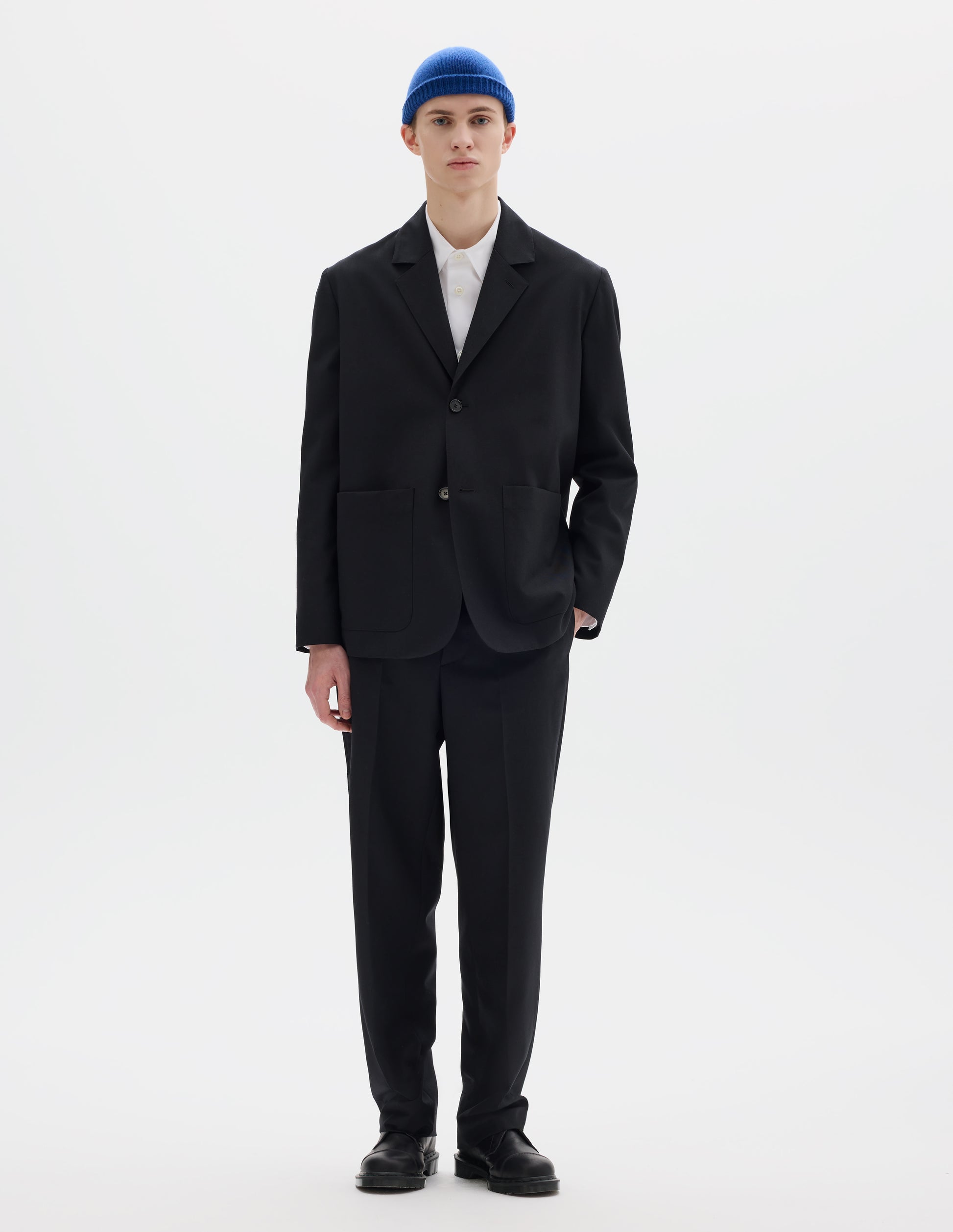 Full body image, front view of model wearing black relaxed wool blazer with dropped shoulders, horn buttons and deep patch pockets. Styled over white shirt, blue hat, matching black trousers and black leather shoes.