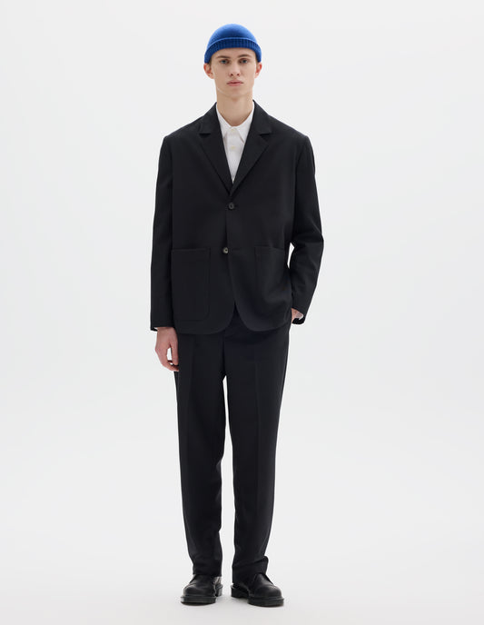 Full body image, front view of model wearing black relaxed wool blazer with dropped shoulders, horn buttons and deep patch pockets. Styled over white shirt, blue hat, matching black trousers and black leather shoes.