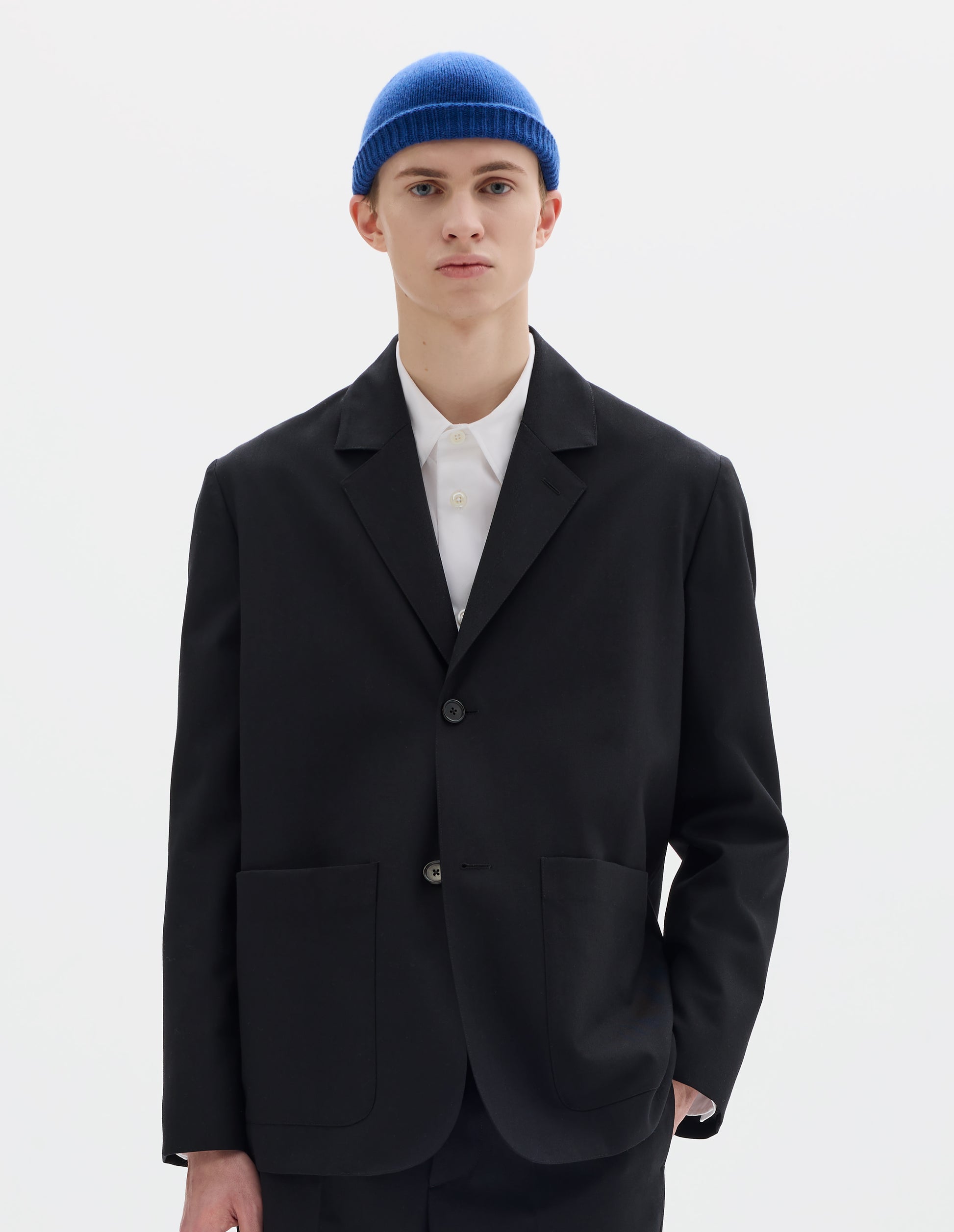 Cropped body image, front view of model wearing black relaxed wool blazer with dropped shoulders, horn buttons and deep patch pockets. Styled over white shirt, blue hat and with matching black trousers.