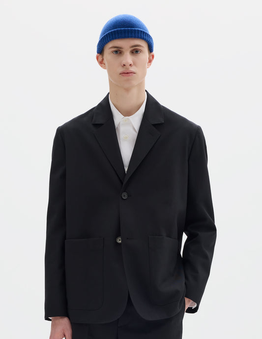 Cropped body image, front view of model wearing black relaxed wool blazer with dropped shoulders, horn buttons and deep patch pockets. Styled over white shirt, blue hat and with matching black trousers.