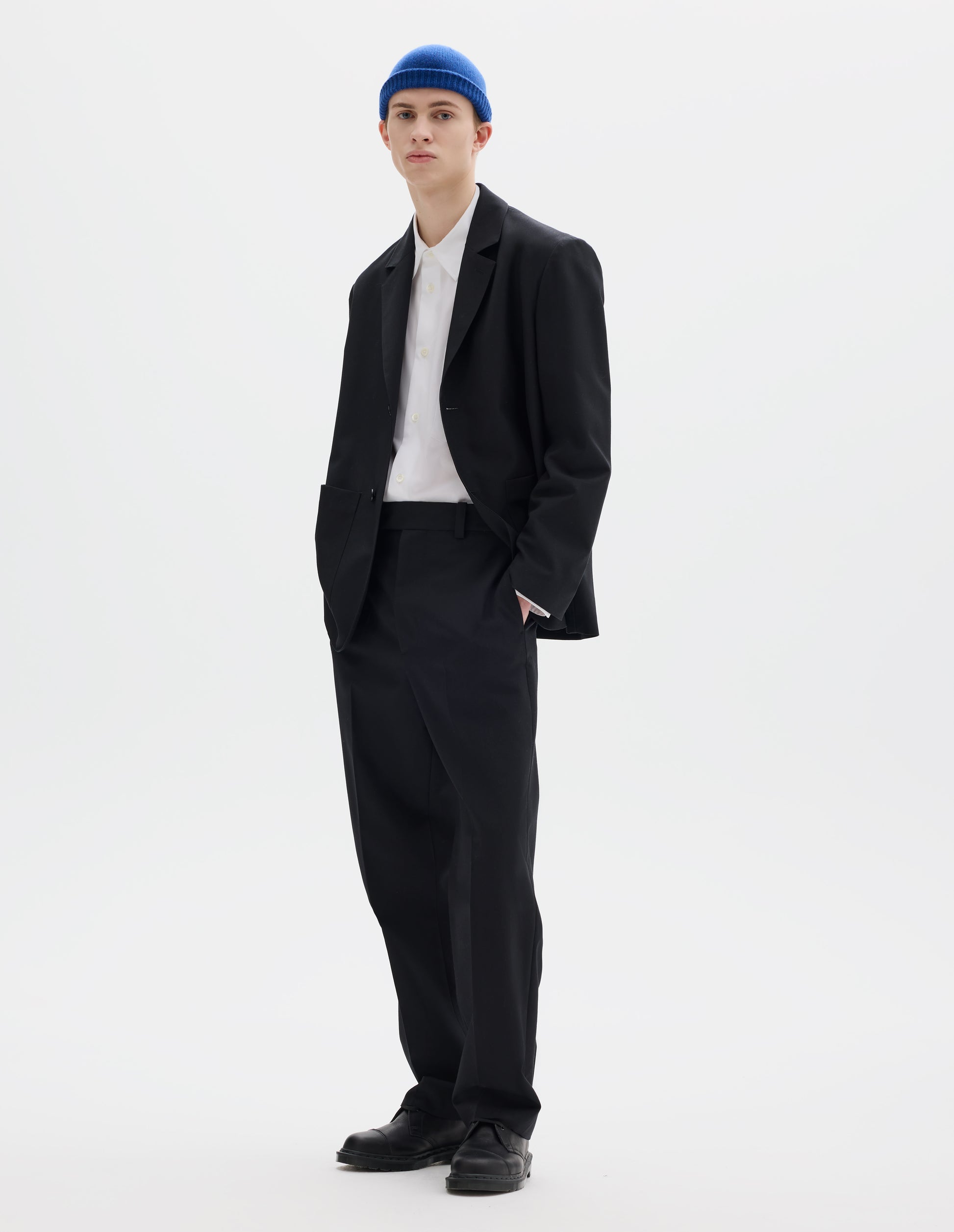Full body image, front view of model wearing relaxed black wool trousers with tapered leg, belt loops and waistband extension with hook and bar fastening. Styled with blue hat, matching black blazer, white shirt and black leather shoes.