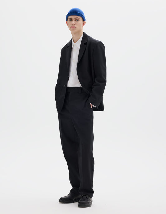 Full body image, front view of model wearing relaxed black wool trousers with tapered leg, belt loops and waistband extension with hook and bar fastening. Styled with blue hat, matching black blazer, white shirt and black leather shoes.