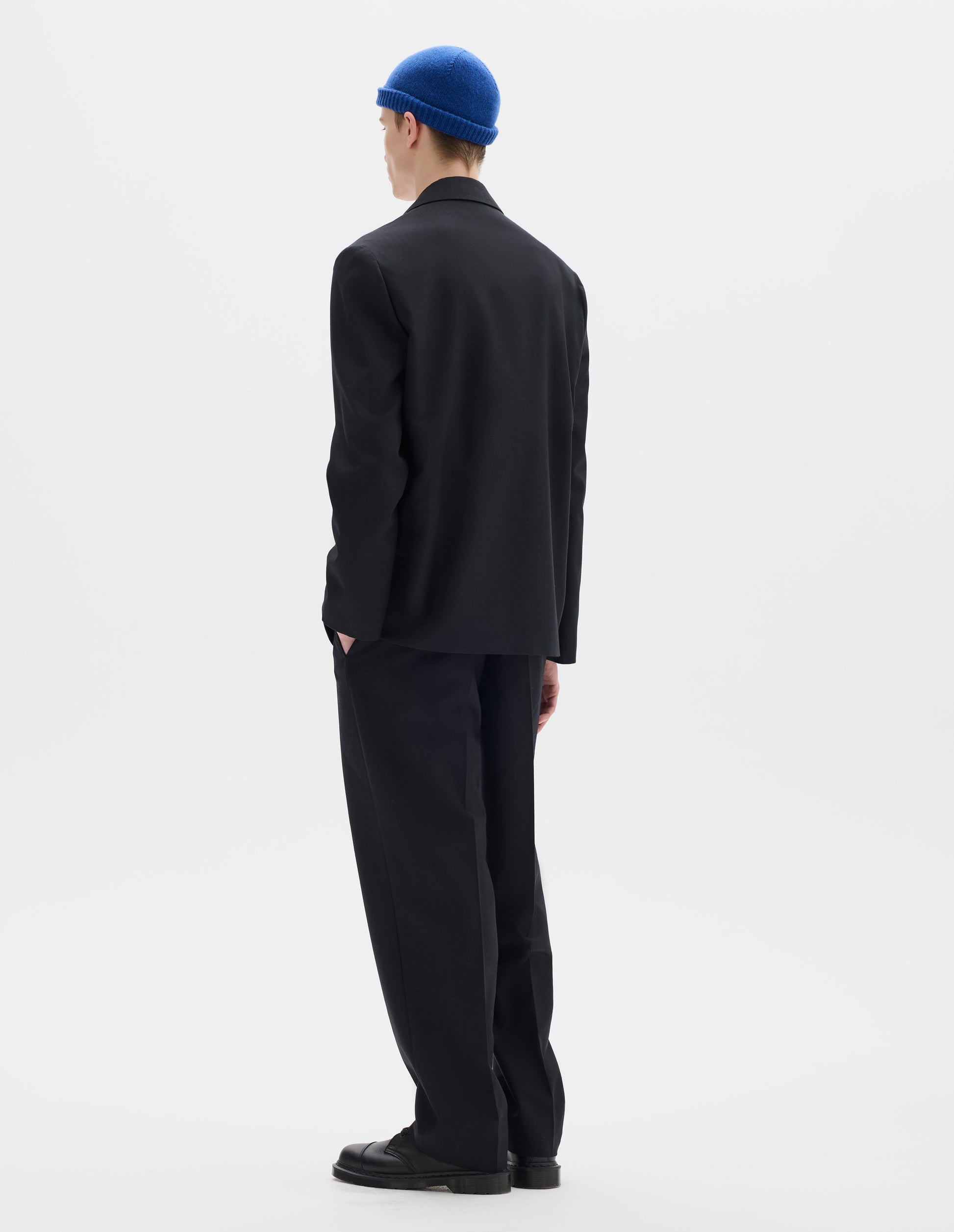 Full body image, back view of model wearing relaxed black wool trousers with tapered leg, belt loops, and with one jetted flap back pocket and one horn button through jetted back pocket. Styled with blue hat, matching black blazer, white shirt and black leather shoes.