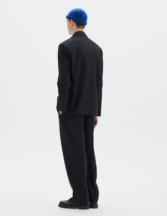 Full body image, back view of model wearing relaxed black wool trousers with tapered leg, belt loops, and with one jetted flap back pocket and one horn button through jetted back pocket. Styled with blue hat, matching black blazer, white shirt and black leather shoes.