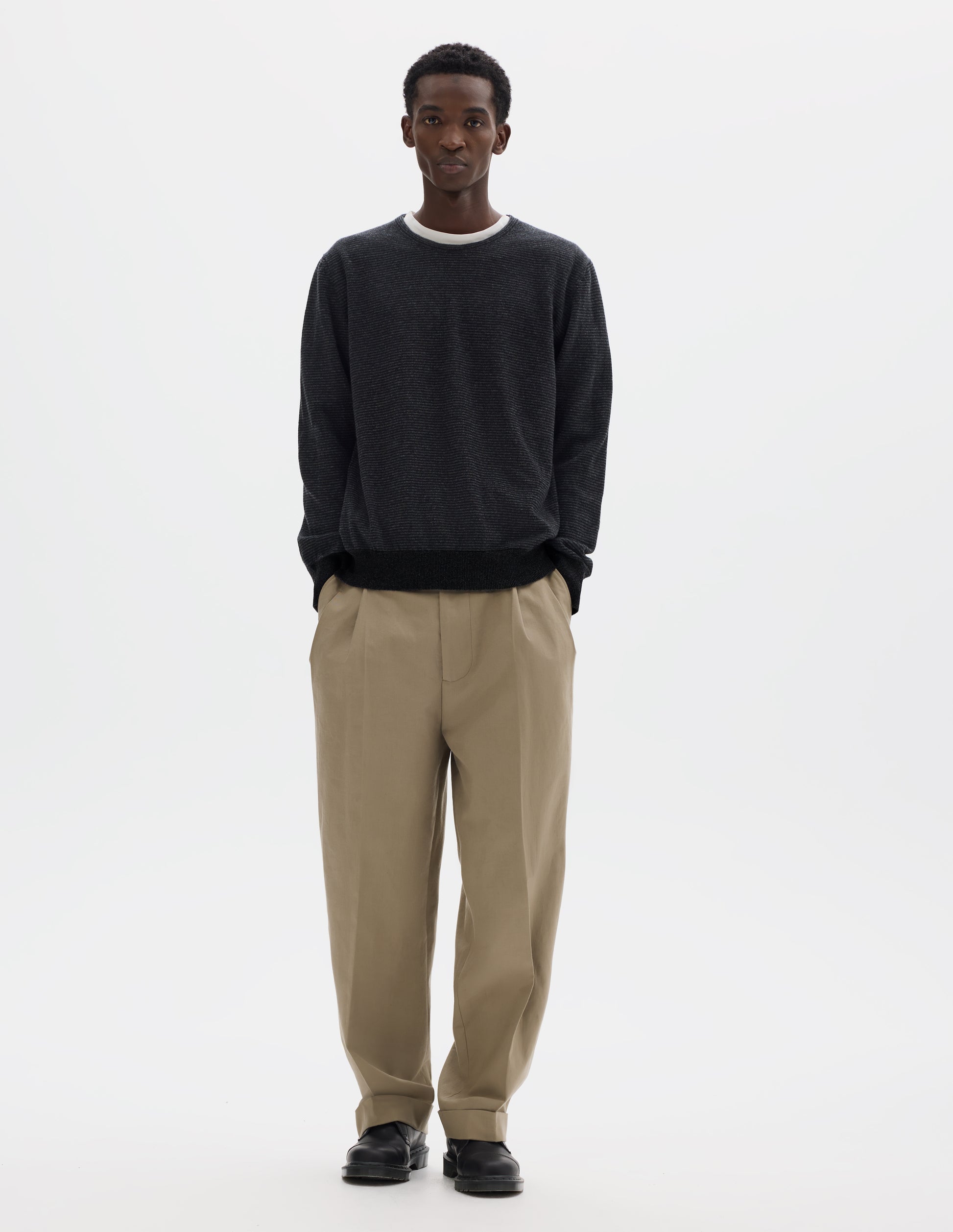 Full body image, front view of model wearing dark grey merino cashmere jumper with black stripes, crew neck with tubular collar, and with contrast rib on cuffs and hem. Styled over white t-shirt and with beige trousers and black leather shoes.