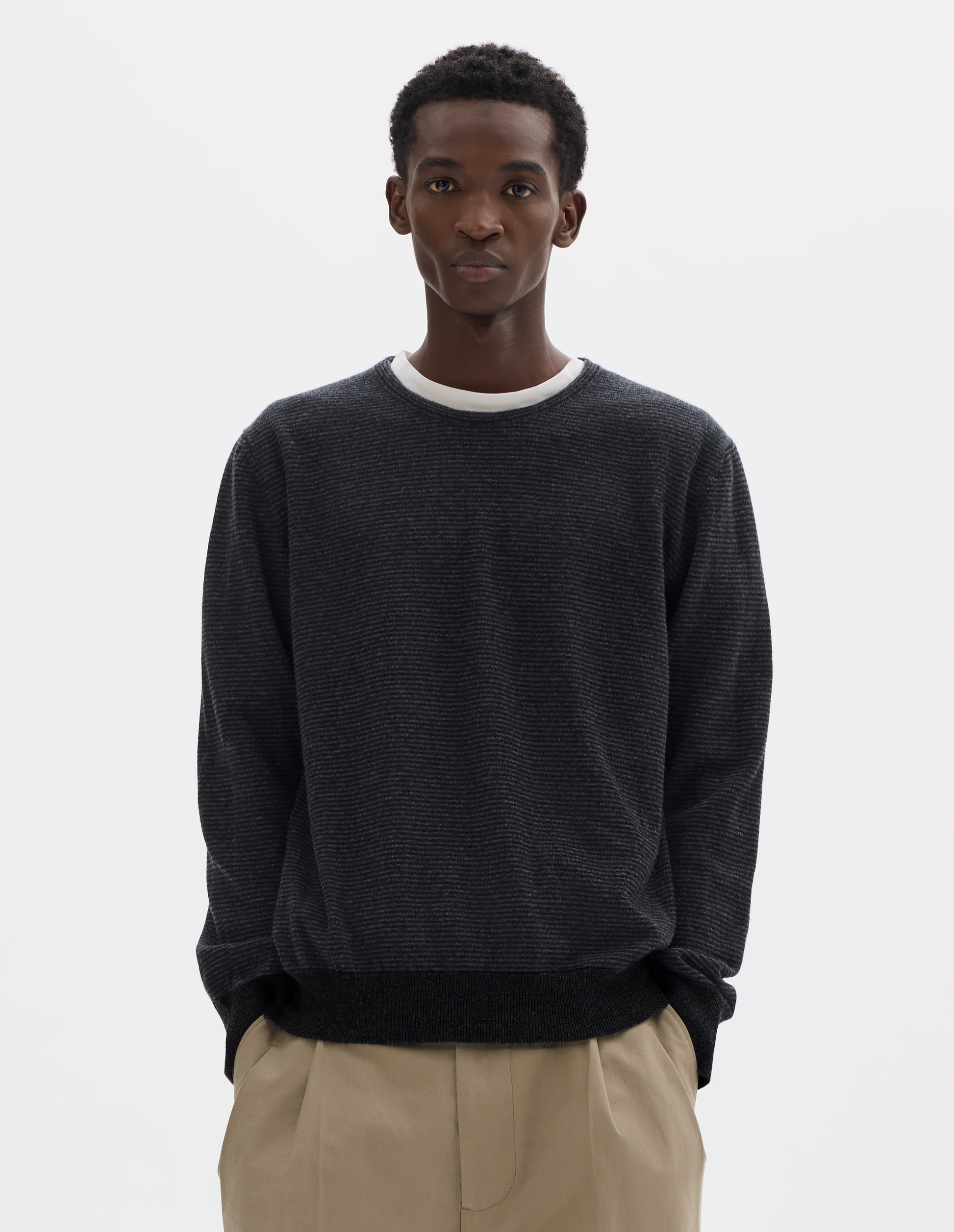 Cropped body image, front view of model wearing dark grey merino cashmere jumper with black stripes, crew neck with tubular collar, and with contrast rib on cuffs and hem. Styled over white t-shirt and with beige trousers.