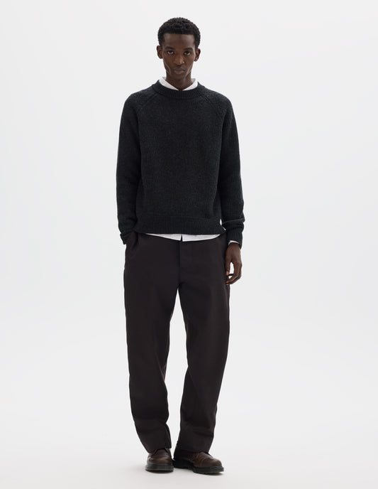 Full body image, front view of model wearing dark grey wool jumper with raglan sleeves, tubular collar and rib detailing at hem and cuffs. Styled over white shirt, and with brown trousers and brown leather shoes.