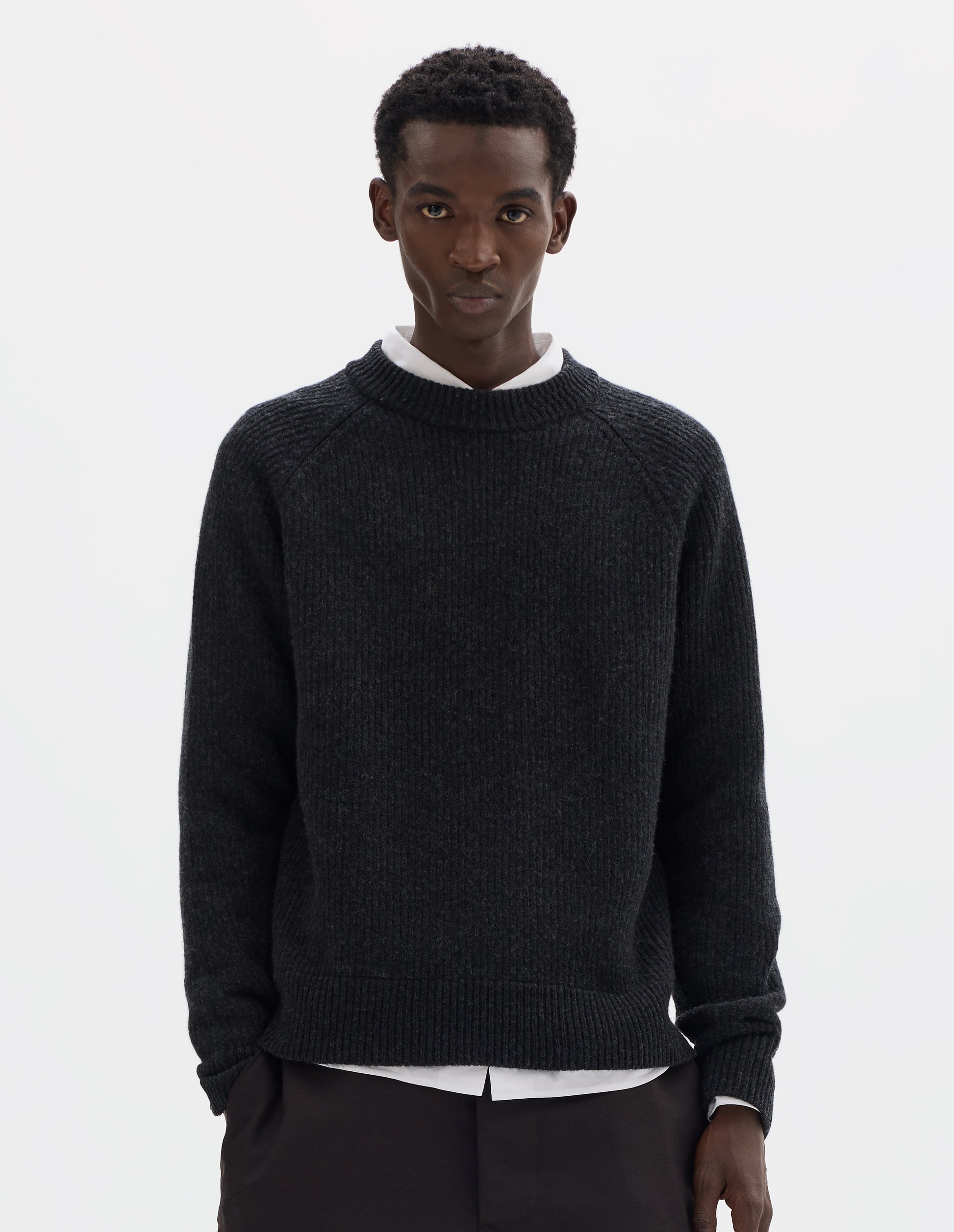 Cropped body image, front view of model wearing dark grey wool jumper with raglan sleeves, tubular collar and rib detailing at hem and cuffs. Styled over white shirt, and with brown trousers.