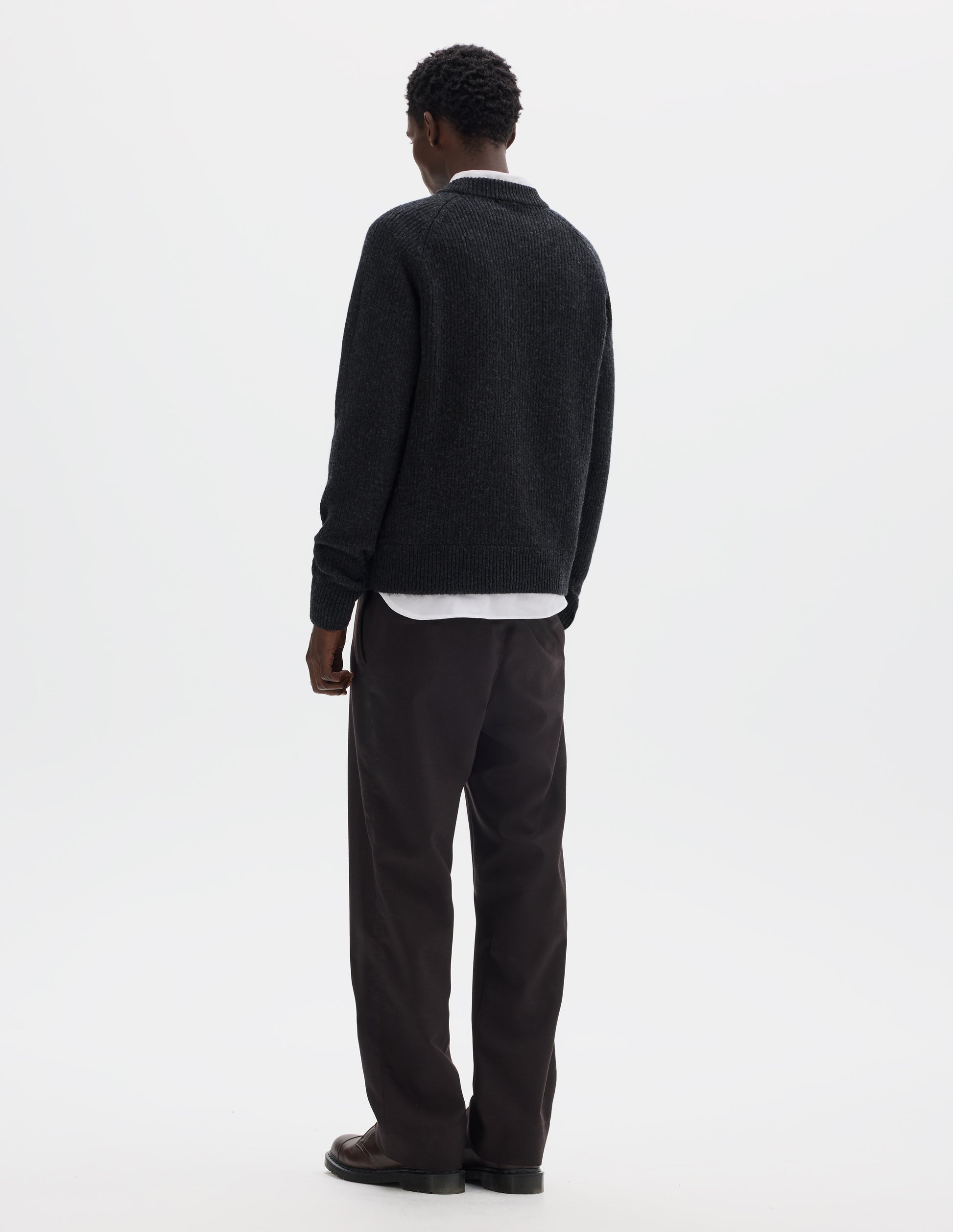 Full body image, back view of model wearing dark grey wool jumper with raglan sleeves, tubular collar and rib detailing at hem and cuffs. Styled over white shirt, and with brown trousers and brown leather shoes.