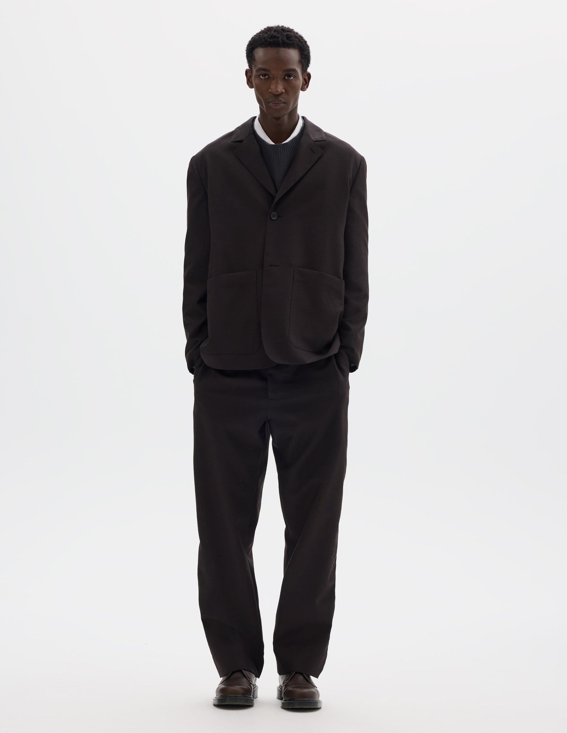 Full body image, front view of model wearing dark brown relaxed wool blazer with dropped shoulders, horn buttons and deep patch pockets. Styled over dark grey jumper and white shirt, with top button closed, and with matching trousers and brown leather shoes.