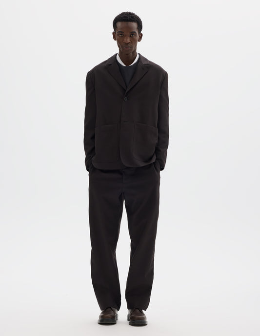 Full body image, front view of model wearing dark brown relaxed wool blazer with dropped shoulders, horn buttons and deep patch pockets. Styled over dark grey jumper and white shirt, with top button closed, and with matching trousers and brown leather shoes.