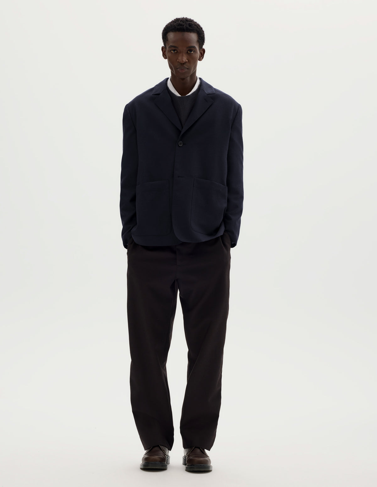 Full body image, front view of model wearing dark blue relaxed wool cotton blazer with dropped shoulders, horn buttons and deep patch pockets. Styled with top button done up over grey jumper and white shirt, and with brown trousers and brown leather shoes.