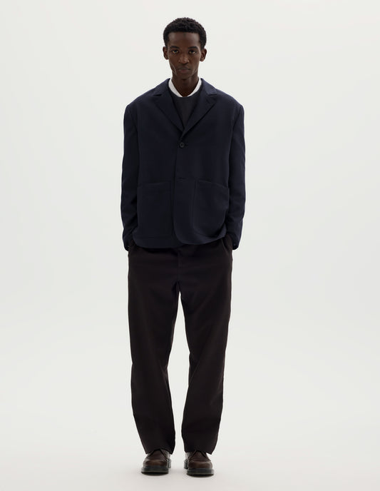 Full body image, front view of model wearing dark blue relaxed wool cotton blazer with dropped shoulders, horn buttons and deep patch pockets. Styled with top button done up over grey jumper and white shirt, and with brown trousers and brown leather shoes.