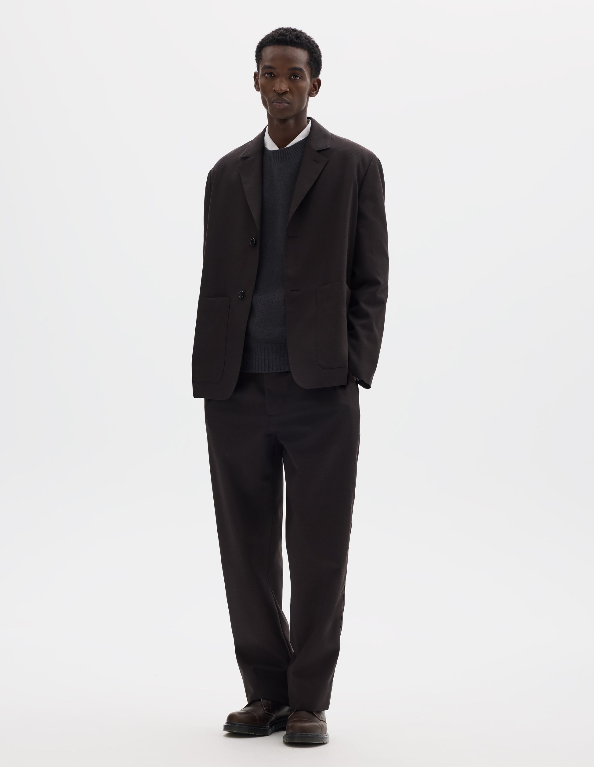 Full body image, front view of model wearing dark brown relaxed wool blazer with dropped shoulders, horn buttons and deep patch pockets. Styled over dark grey jumper and white shirt, and with matching trousers and brown leather shoes.
