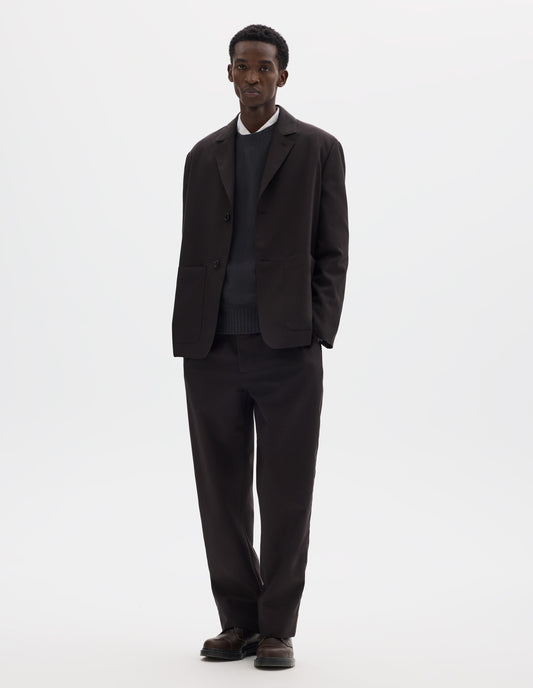Full body image, front view of model wearing dark brown relaxed wool blazer with dropped shoulders, horn buttons and deep patch pockets. Styled over dark grey jumper and white shirt, and with matching trousers and brown leather shoes.