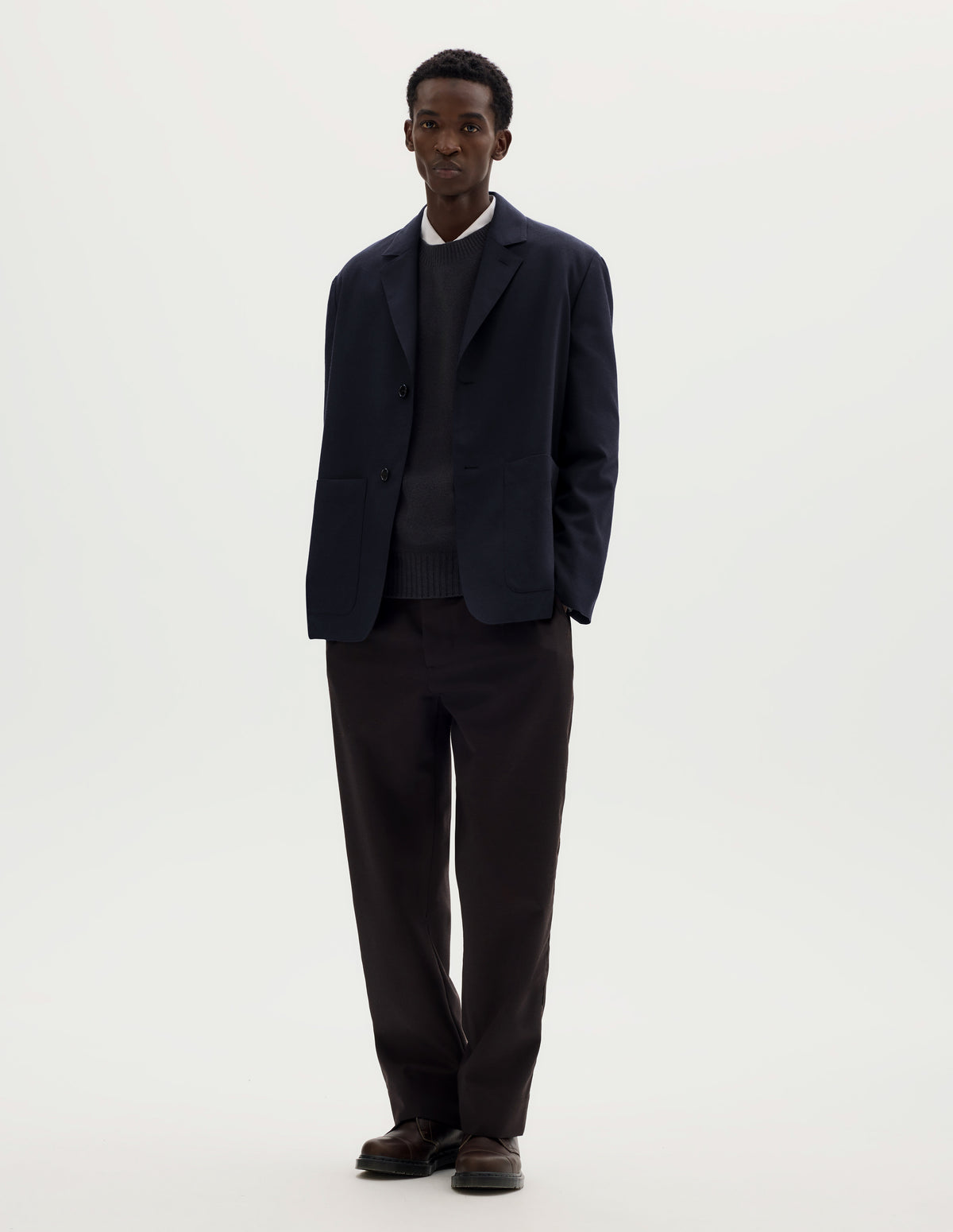 Full body image, front view of model wearing dark blue relaxed wool cotton blazer with dropped shoulders, horn buttons and deep patch pockets. Styled over grey jumper and white shirt, and with brown trousers and brown leather shoes.