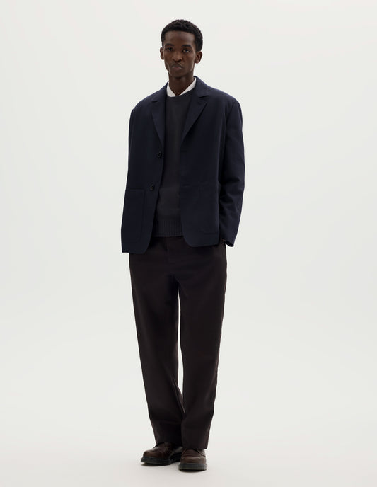Full body image, front view of model wearing dark blue relaxed wool cotton blazer with dropped shoulders, horn buttons and deep patch pockets. Styled over grey jumper and white shirt, and with brown trousers and brown leather shoes.