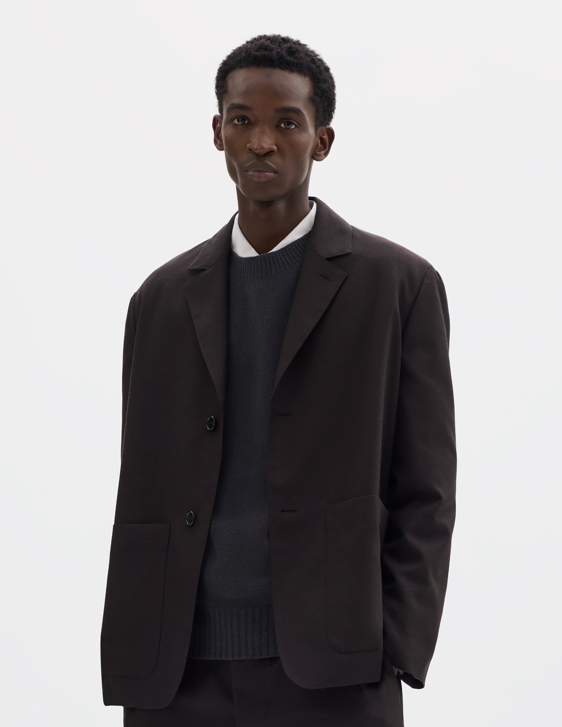 Cropped body image, front view of model wearing dark brown relaxed wool blazer with dropped shoulders, horn buttons and deep patch pockets. Styled over dark grey jumper and white shirt, and with matching trousers.