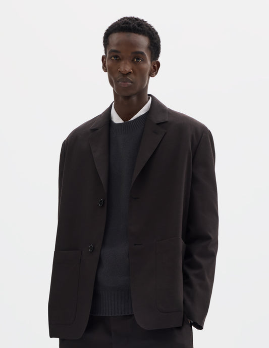 Cropped body image, front view of model wearing dark brown relaxed wool blazer with dropped shoulders, horn buttons and deep patch pockets. Styled over dark grey jumper and white shirt, and with matching trousers.