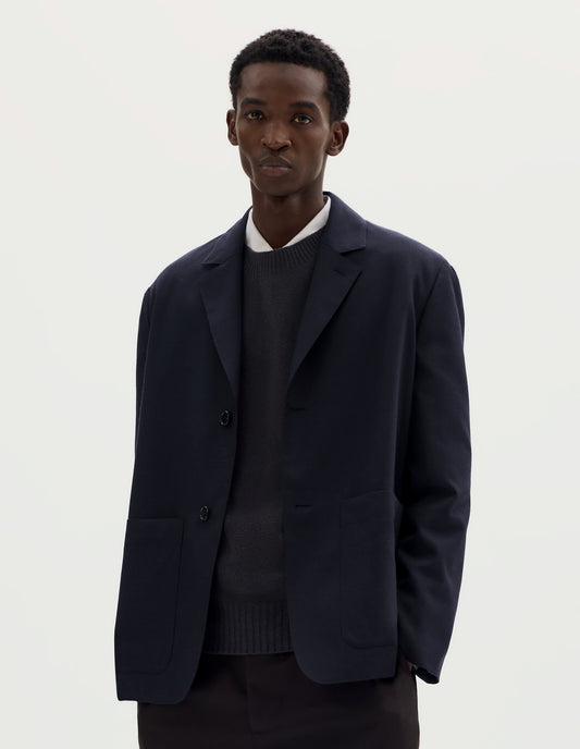 Cropped body image, front view of model wearing dark blue relaxed wool cotton blazer with dropped shoulders, horn buttons and deep patch pockets. Styled over grey jumper and white shirt, and with brown trousers.