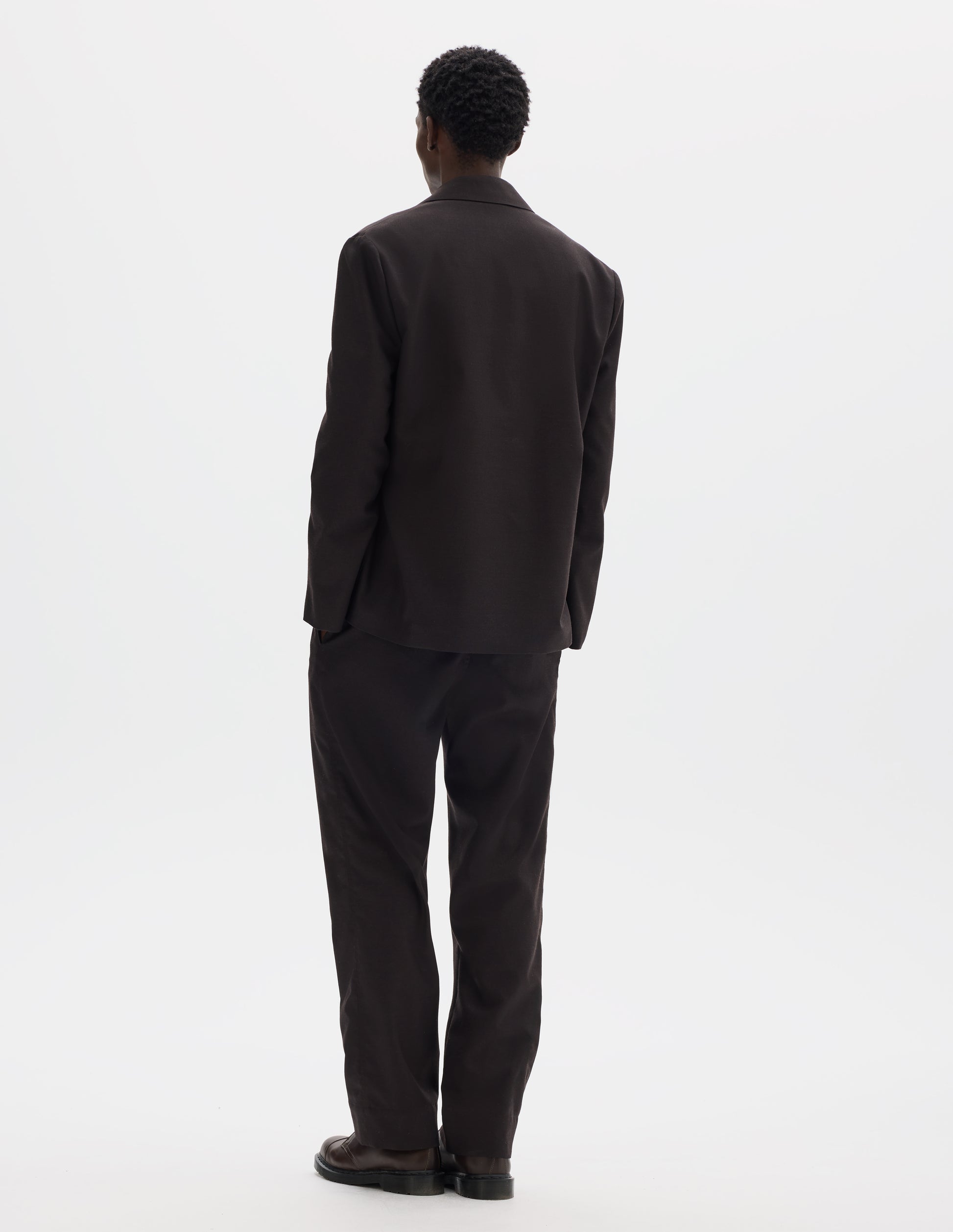 Full body image, back view of model wearing dark brown relaxed wool blazer with dropped shoulders. Styled with matching trousers and brown leather shoes.