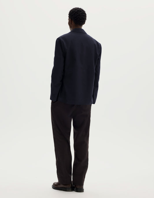 Full body image, back view of model wearing dark blue relaxed wool cotton blazer with dropped shoulders. Styled with brown trousers and brown leather shoes.