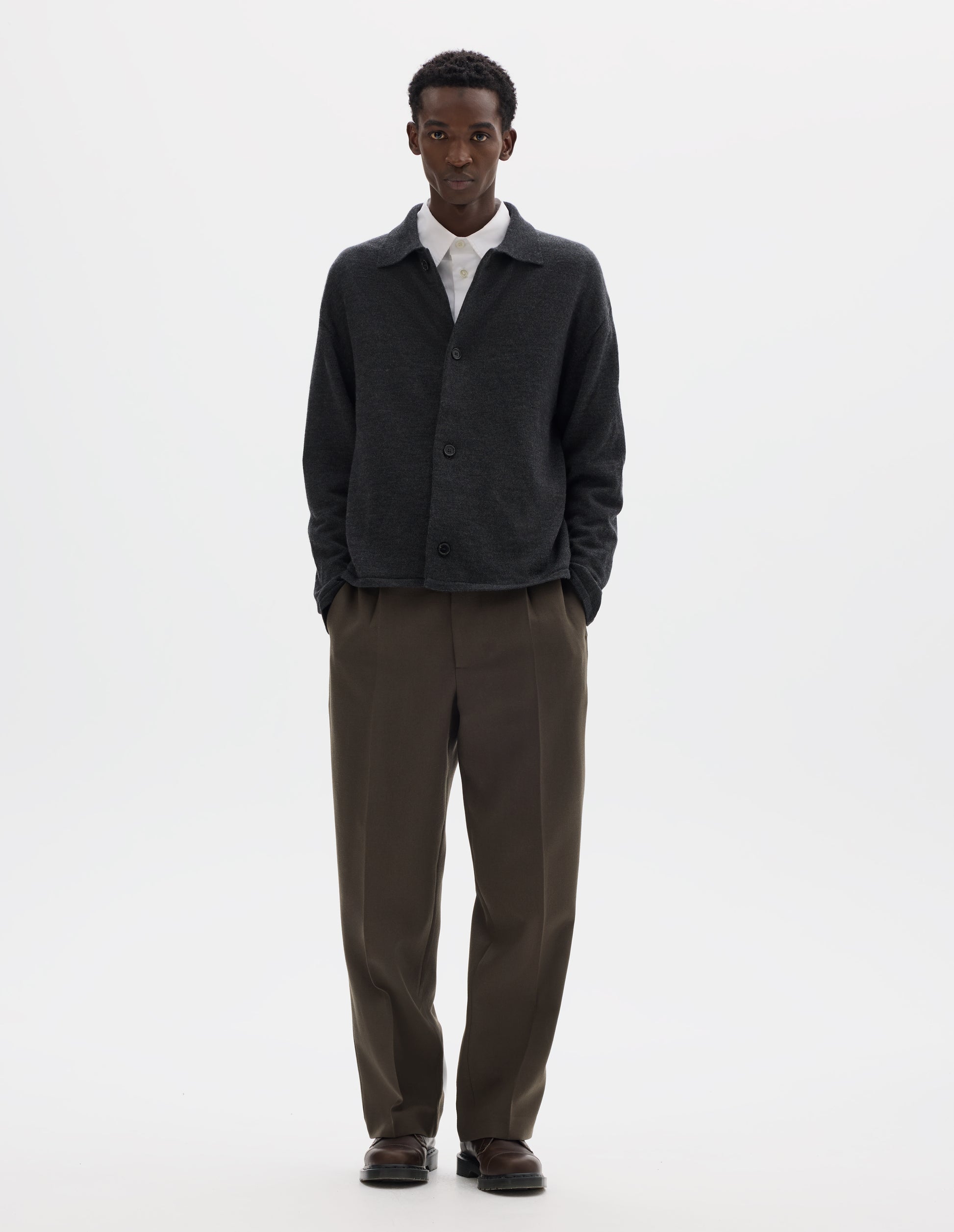 Full body image, front view of model wearing relaxed dark grey merino cardigan with jersey stitch, fully fashioned collar, horn button fastening, set-in fully fashioned sleeves, and tubular hem and cuffs. Styled with top button undone over white shirt, with  brown trousers and brown leather shoes.