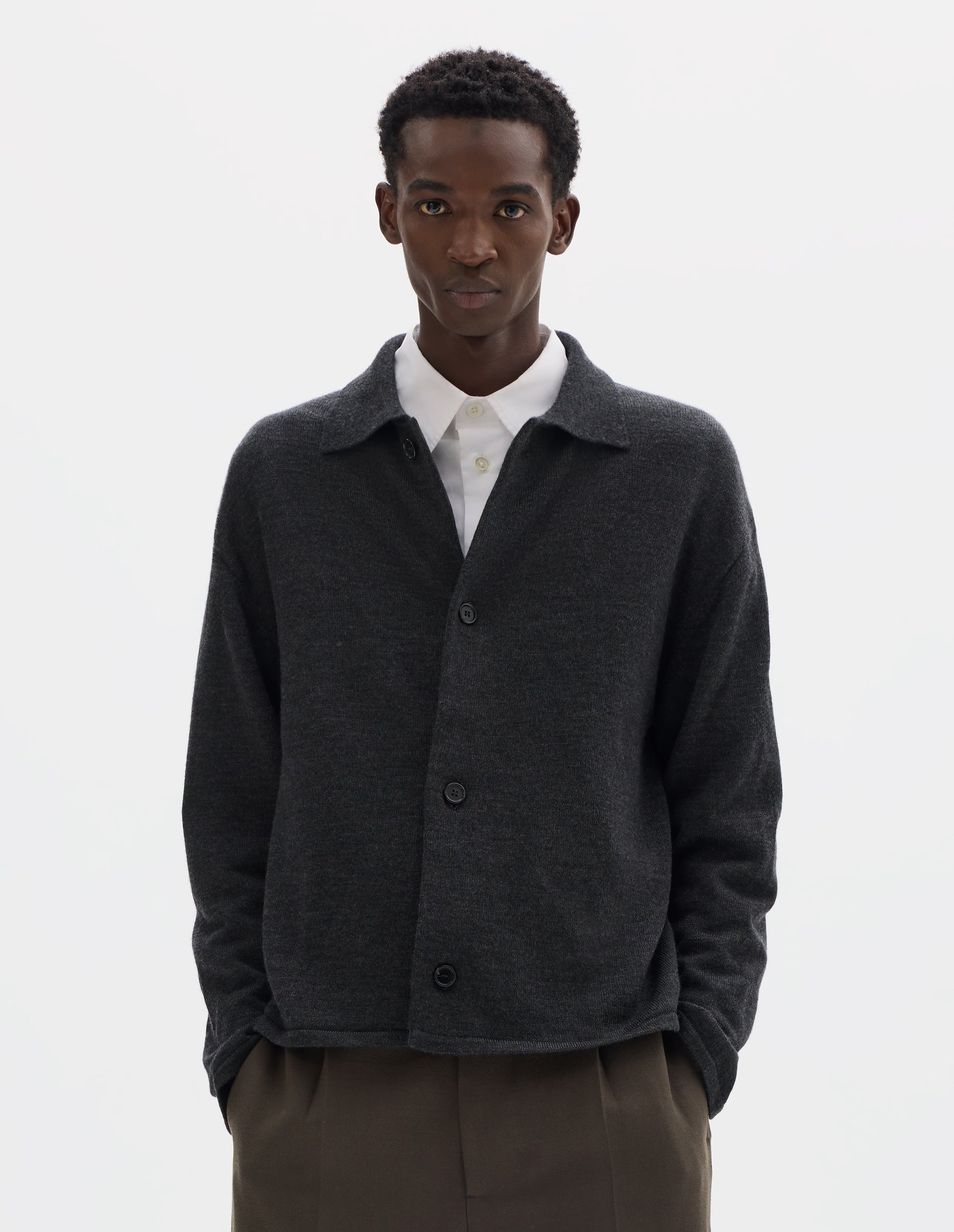 Cropped body image, front view of model wearing relaxed dark grey merino cardigan with jersey stitch, fully fashioned collar, horn button fastening, set-in fully fashioned sleeves, and tubular hem and cuffs. Styled with top button undone over white shirt and with  brown trousers.