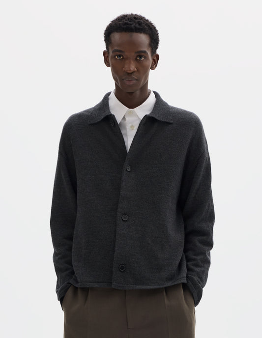 Cropped body image, front view of model wearing relaxed dark grey merino cardigan with jersey stitch, fully fashioned collar, horn button fastening, set-in fully fashioned sleeves, and tubular hem and cuffs. Styled with top button undone over white shirt and with  brown trousers.