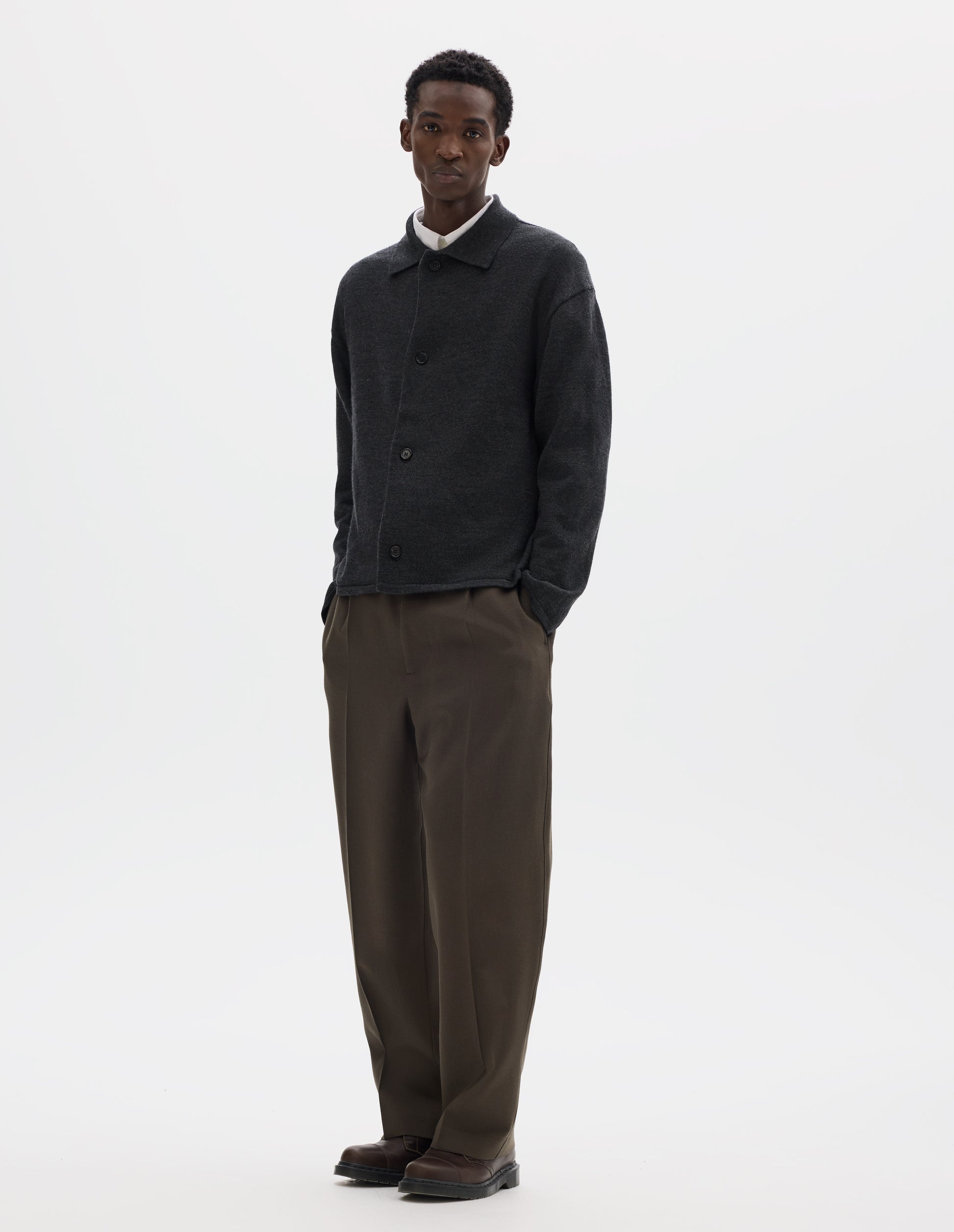 Full body image, front view of model wearing relaxed dark grey merino cardigan with jersey stitch, fully fashioned collar, horn button fastening, set-in fully fashioned sleeves, and tubular hem and cuffs. Styled with buttons closed over white shirt, with brown trousers and brown leather shoes.