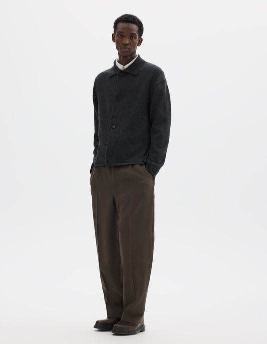 Full body image, front view of model wearing relaxed dark grey merino cardigan with jersey stitch, fully fashioned collar, horn button fastening, set-in fully fashioned sleeves, and tubular hem and cuffs. Styled with buttons closed over white shirt, with brown trousers and brown leather shoes.