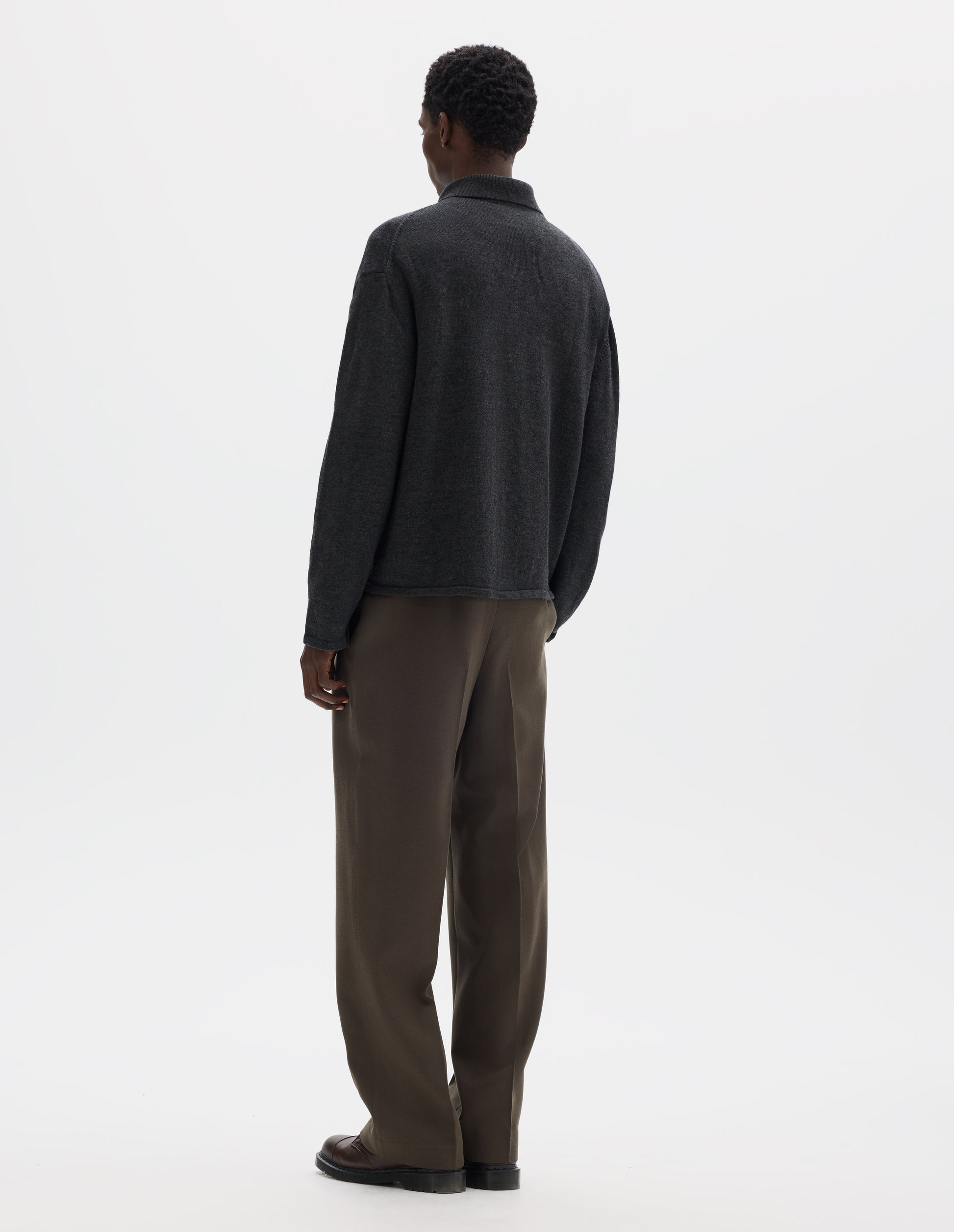 Full body image, back view of model wearing relaxed dark  grey merino cardigan with jersey stitch, fully fashioned collar, set-in fully fashioned sleeves, and tubular hem and cuffs. Styled with brown trousers and brown leather shoes.
