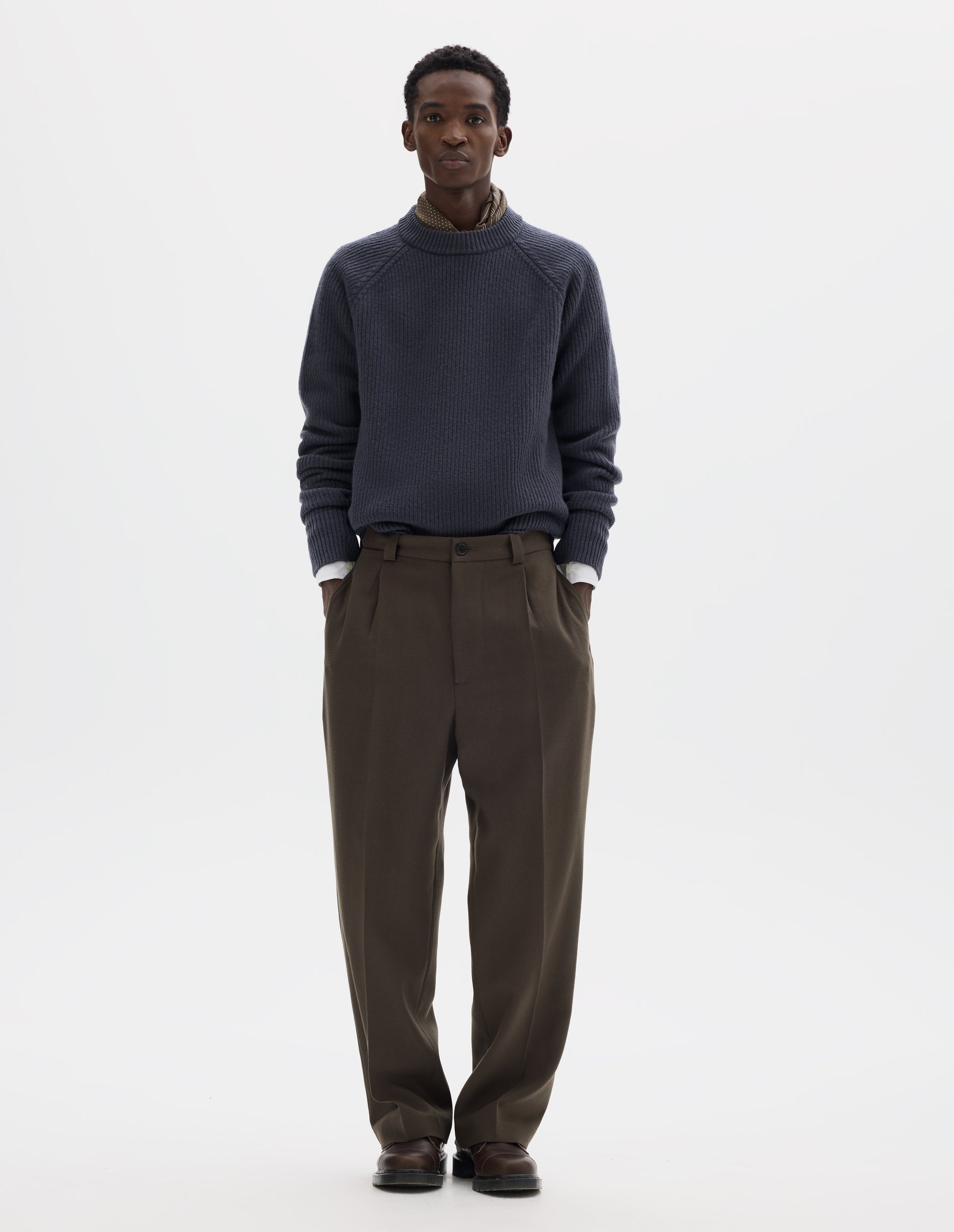 Full body image, front view of model wearing brown straight leg wool trousers with horn button and zip fly fastening, regular belt loops, single pleats, hidden ticket pocket and side pockets. Styled with grey jumper, white shirt, dark beige silk neck scarf and brown leather shoes.