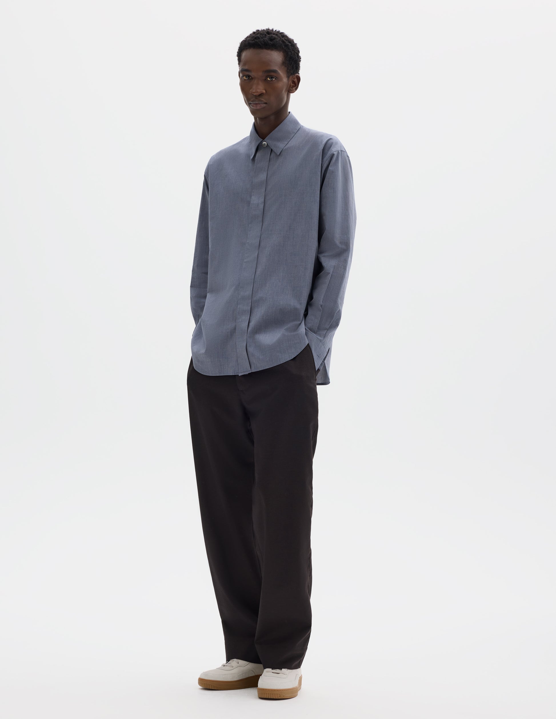 Full body image, front view of model wearing blue cotton shirt with enclosed placket and mother of pearl button on collar, hanger loop on back neck and curved hem. Styled with dark brown trousers and beige nubuck trainers.