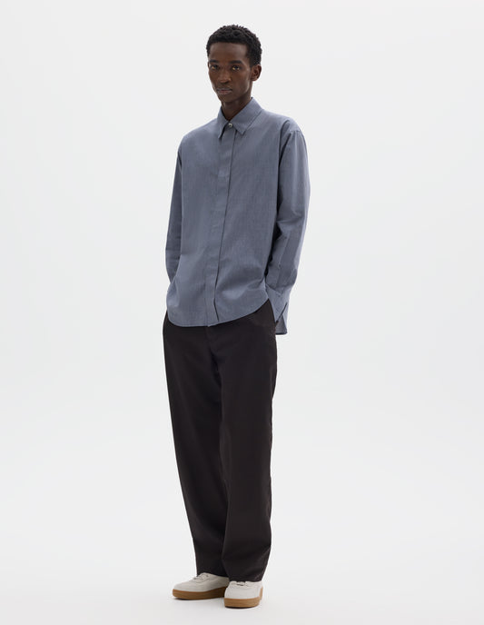 Full body image, front view of model wearing blue cotton shirt with enclosed placket and mother of pearl button on collar, hanger loop on back neck and curved hem. Styled with dark brown trousers and beige nubuck trainers.