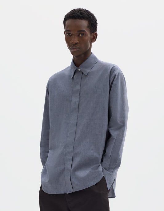 Cropped body image, front view of model wearing blue cotton shirt with enclosed placket and mother of pearl button on collar, hanger loop on back neck and curved hem. Styled with dark brown trousers.