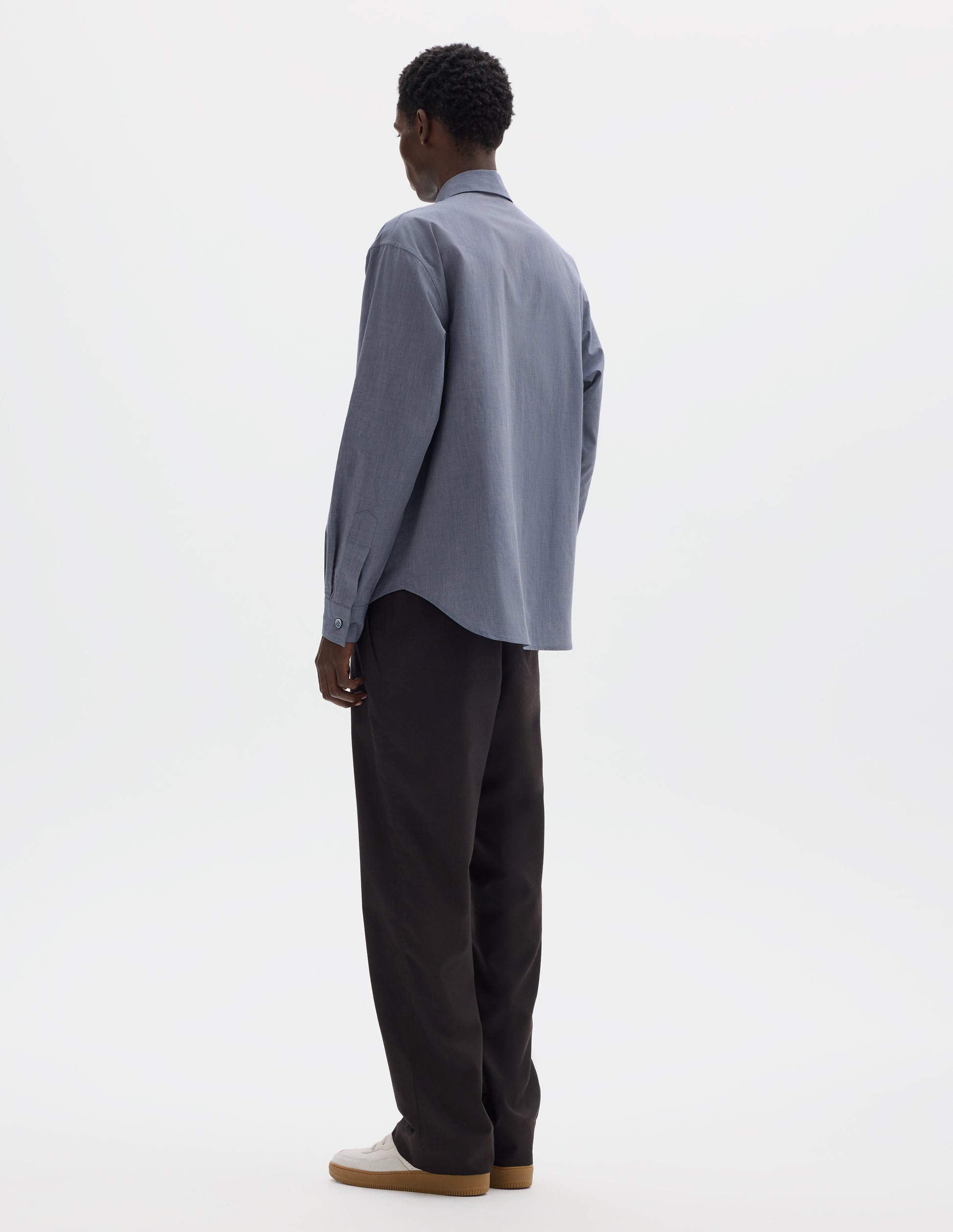 Full body image, back view of model wearing blue cotton shirt with mother of pearl buttons on cuffs and curved hem. Styled with dark brown trousers and beige nubuck trainers.